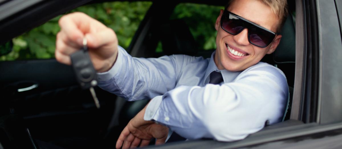 Foto van een lachende jongeman achter het stuur van een auto met een autosleutel in zijn hand