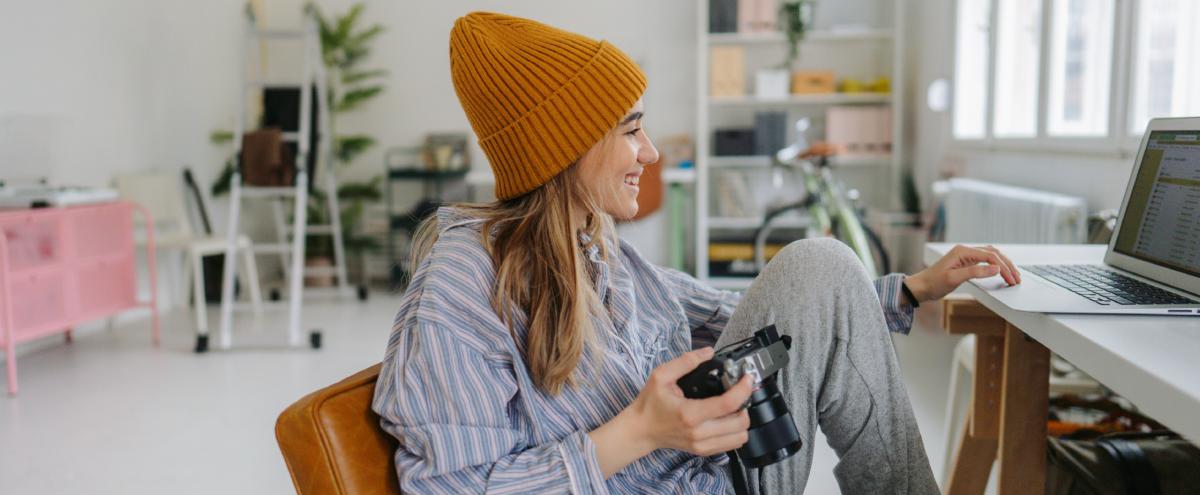 Jonge freelance fotografe zit ontspannen lachend achter een bureau met een fotocamera in haar hand