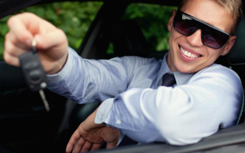 Foto van een lachende jongeman achter het stuur van een auto met een autosleutel in zijn hand